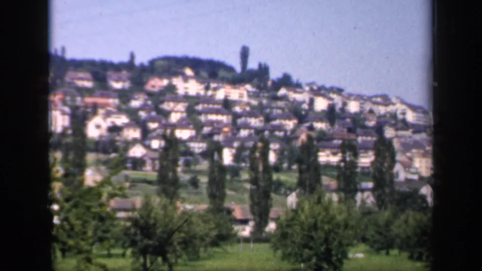 1949 England 8mm frame showing a hillside village with dense houses, slender trees, green fields, and a blue sky.