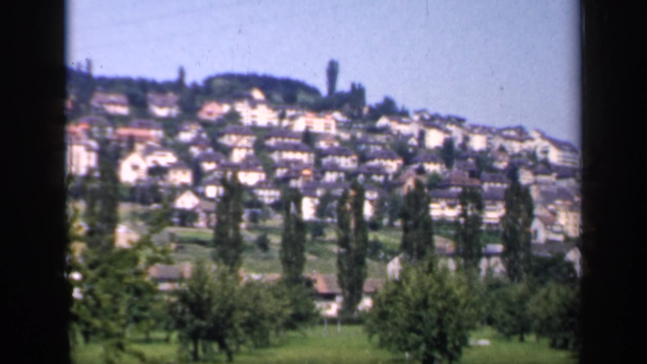 1949 England home-movie frame of hillside houses, green foreground trees, and a clear blue sky with visible 8mm texture.
