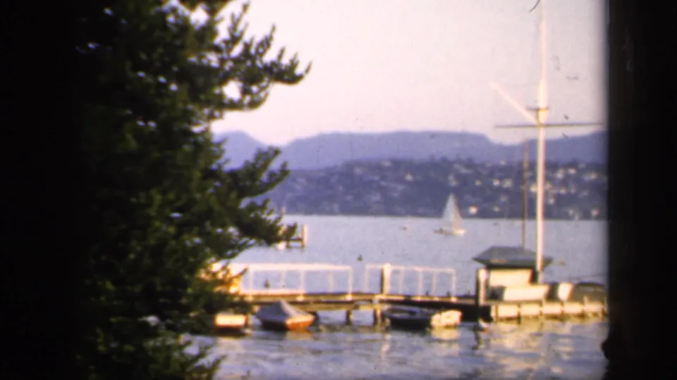 1949 England 8mm frame showing a calm lake, wooden pier, moored boats, a distant sailboat, hillside homes, and low hills beyond the water.