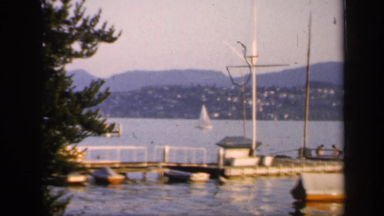1949 England lakeside view with a wooden pier, small boats, sailboat on open water, hillside houses, low hills, and dark film-frame edges.