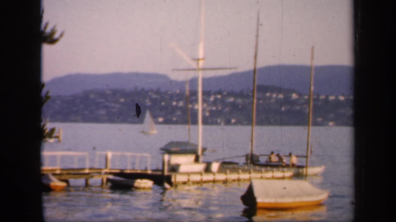 1949 England home-movie frame showing a moored sailboat near a pier, tall masts, calm water, a distant sail, hillside homes, and low hills.