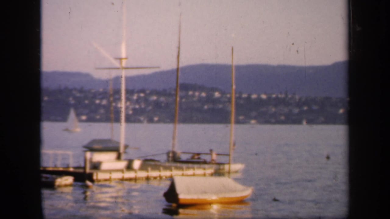 1949 England 8mm frame of a moored sailboat by a pier, possible tiny people aboard, a calm lake, distant sailboat, hillside homes, and low hills.
