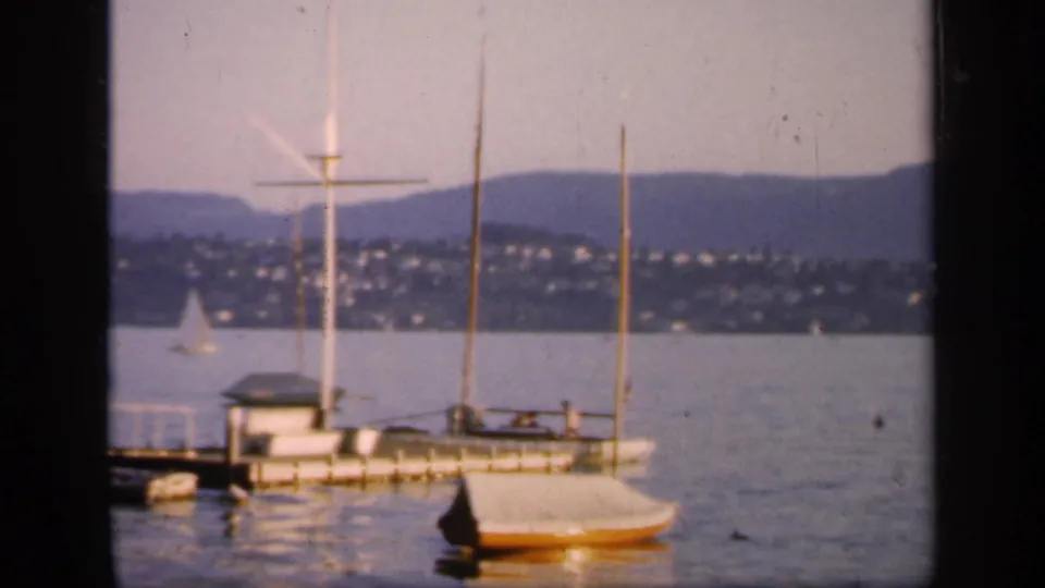 1949 England 8mm frame of a moored sailboat by a pier, possible tiny people aboard, a calm lake, distant sailboat, hillside homes, and low hills.