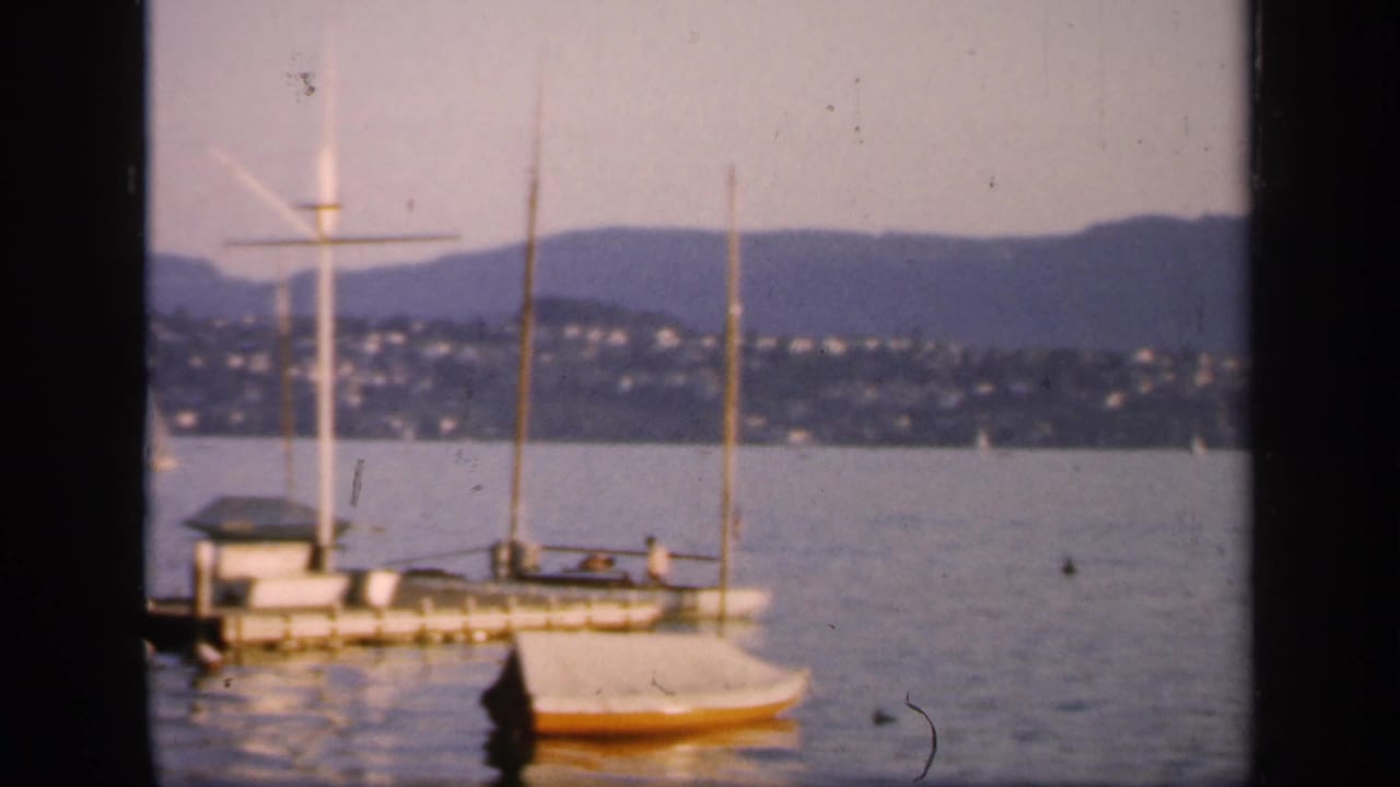 1949 England harbor view with small moored sailboats, a docked vessel, distant hillside houses, and mountains seen through dark window framing.