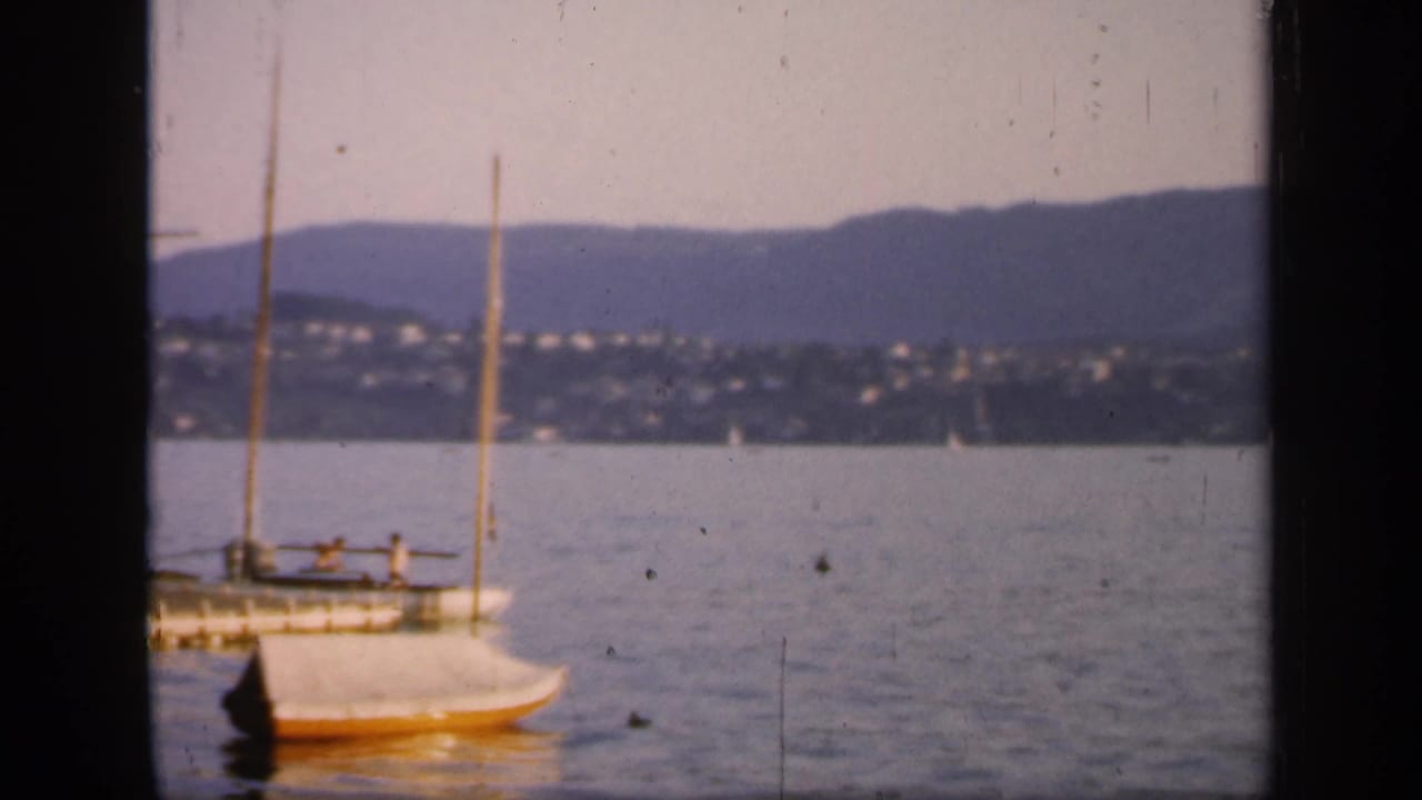A 1949 England harbor frame shows a docked mast vessel and a small sailboat in front of distant shoreline houses and low mountains.