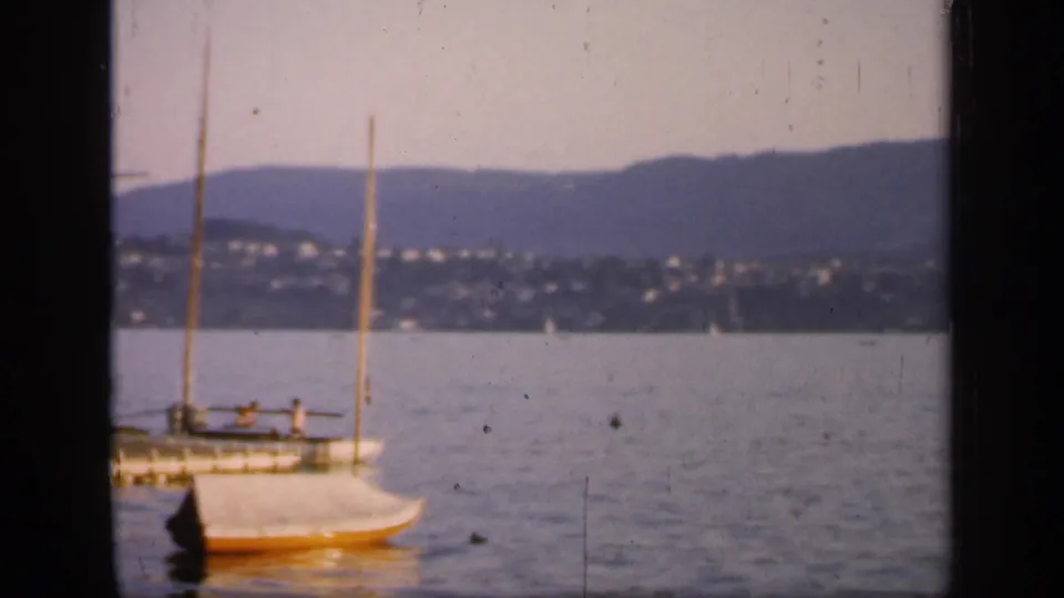 A 1949 England harbor frame shows a docked mast vessel and a small sailboat in front of distant shoreline houses and low mountains.