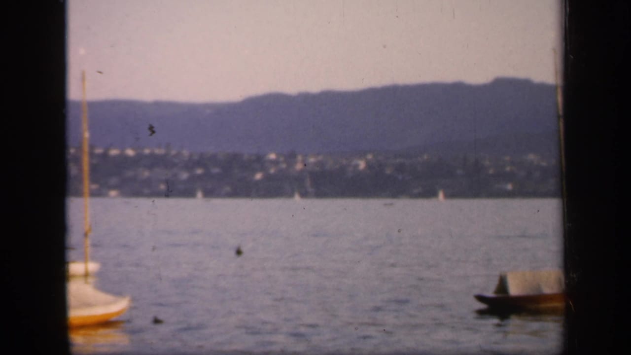 The camera has shifted across the 1949 England harbor, leaving boats partly cropped against water, hillside settlement, and mountain backdrop.