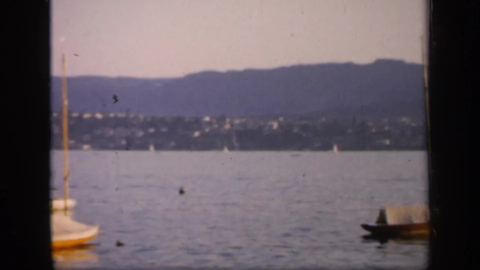 The camera has shifted across the 1949 England harbor, leaving boats partly cropped against water, hillside settlement, and mountain backdrop.