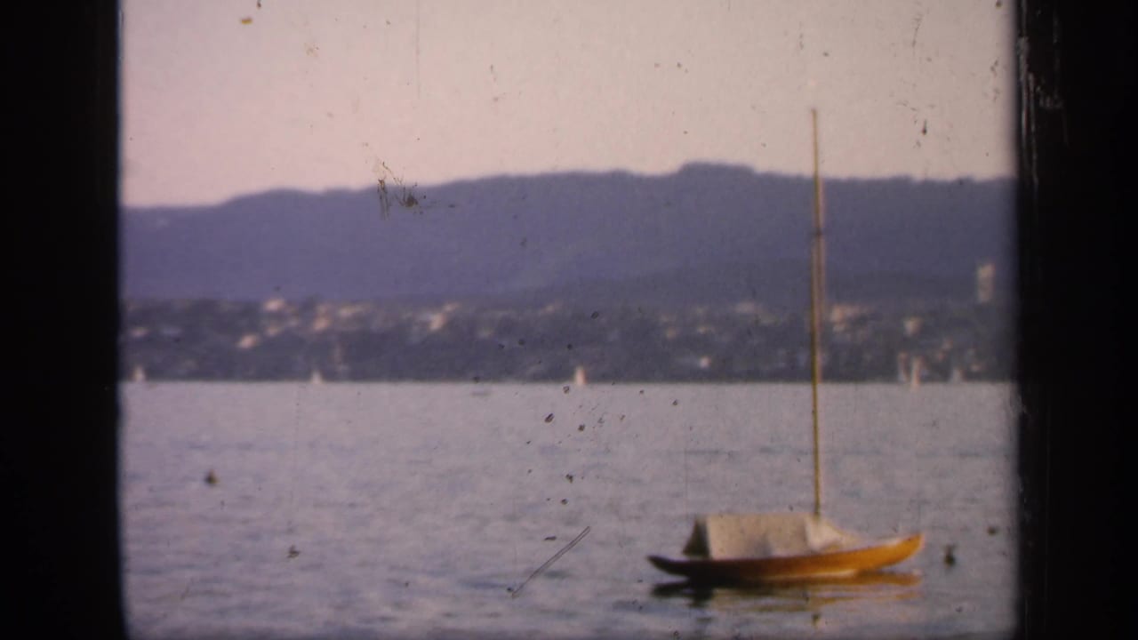 A small moored sailboat sits on the harbor in 1949 England footage, framed by distant shoreline houses, low mountains, and visible 8mm dust.