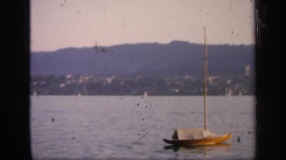 A small moored sailboat sits on the harbor in 1949 England footage, framed by distant shoreline houses, low mountains, and visible 8mm dust.