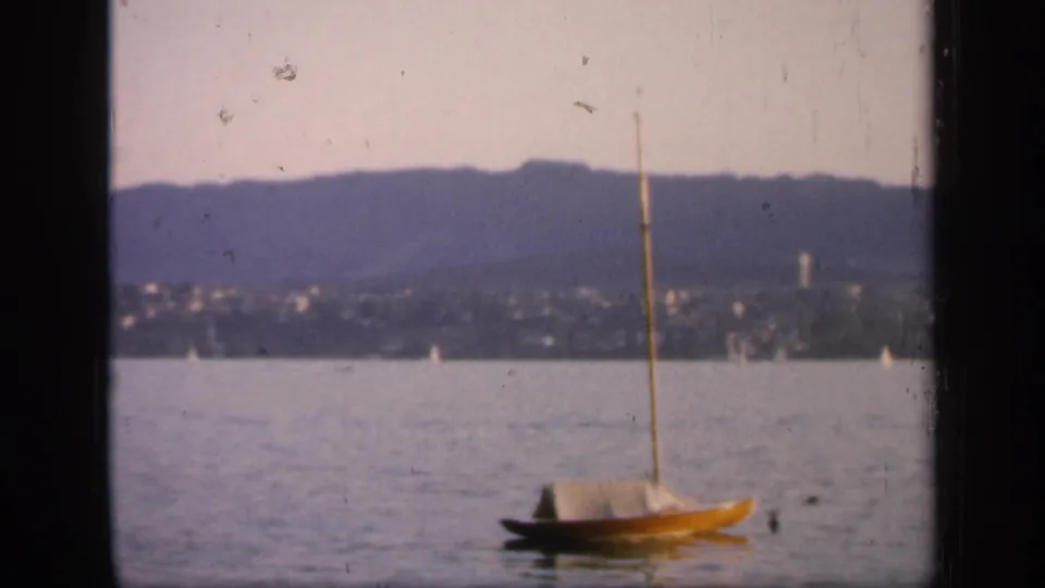 1949 England 8mm frame of a small yellow sailboat on calm water, with hills, shoreline buildings, and dark foreground borders.