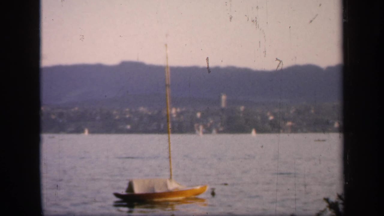 Small sailboat centered on calm water with a town and hills in soft focus behind it.