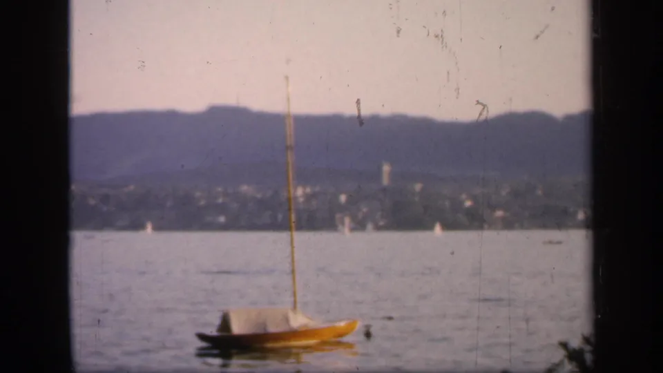Small yellow sailboat on an English waterway in 1949, framed by dark edges with distant town, hills, and tiny sails beyond.