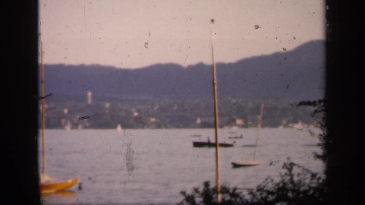 End frame of the 1949 England sailing scene, with the yellow boat partly cropped, distant boats on calm water, and hills behind.