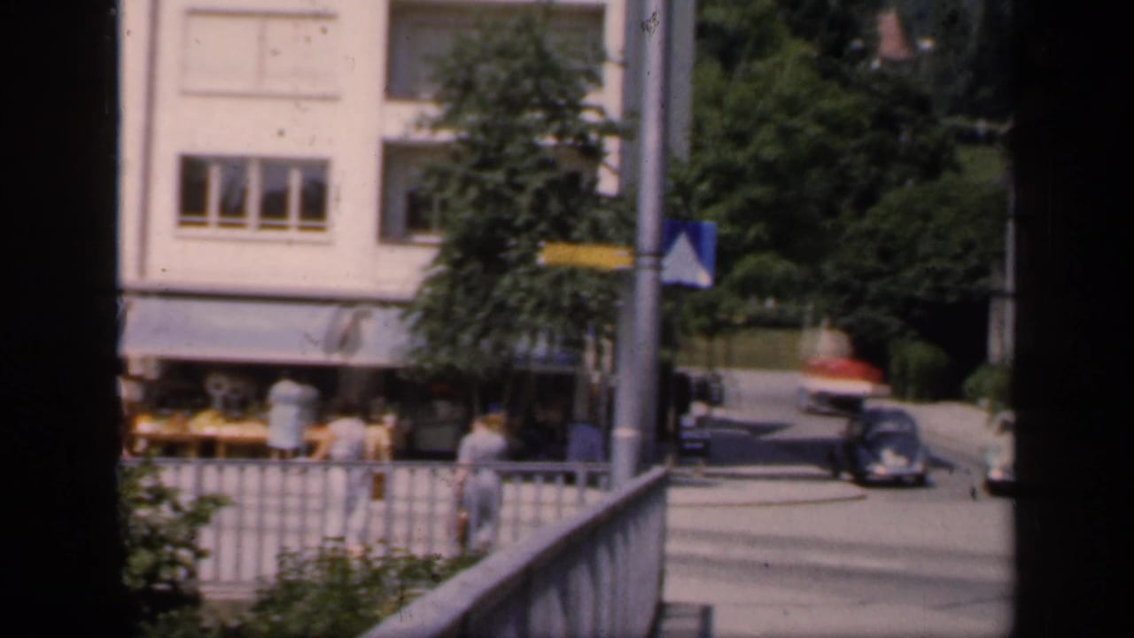1949 English home-movie view across a town street with pedestrians, railings, road signs, and mid-century cars near pale buildings.