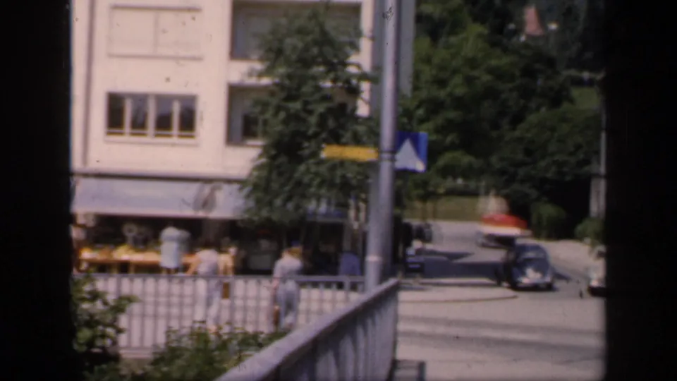 1949 English home-movie view across a town street with pedestrians, railings, road signs, and mid-century cars near pale buildings.