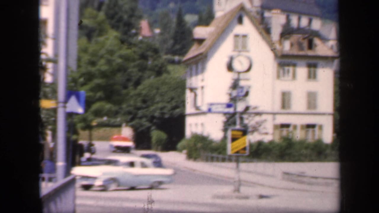 Cars pass near a pale building with a clock and signs, with trees and hills behind.