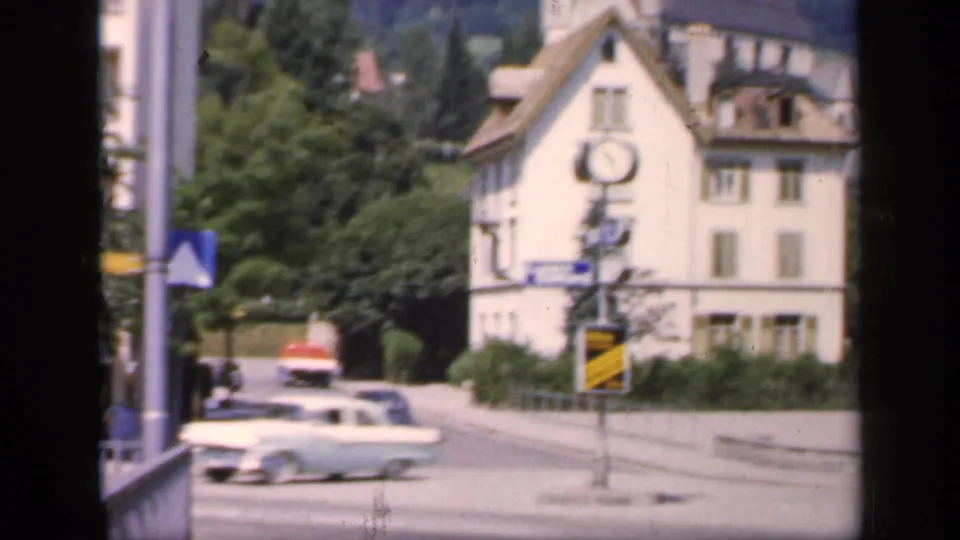 1949 English town view with classic cars near a clock-fronted building, street signs, trees, and wooded hills in the background.