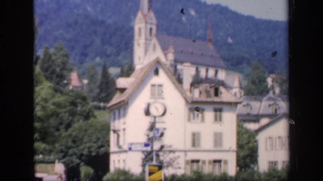 1949 English home-movie frame showing a pale building with a clock below a church steeple, with trees, rooftops, and hills beyond.