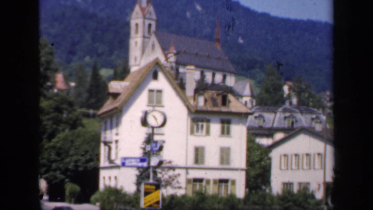 1949 English town frame with a clock-fronted building, church steeple, nearby rooftops, road signage, trees, and wooded hills.