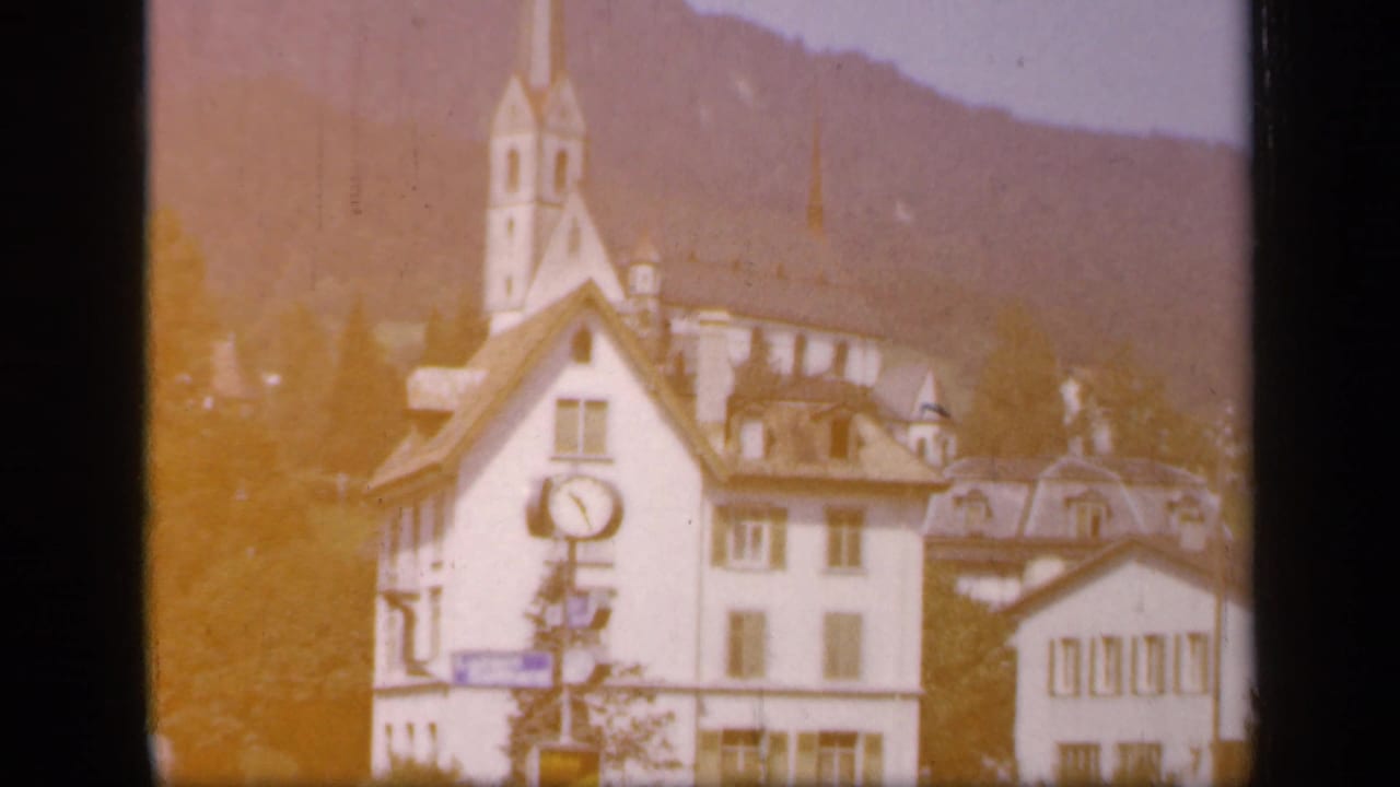 1949 home-movie frame of a white clock-fronted building before a church spire, with hills behind and warm exposure flare at the edge.