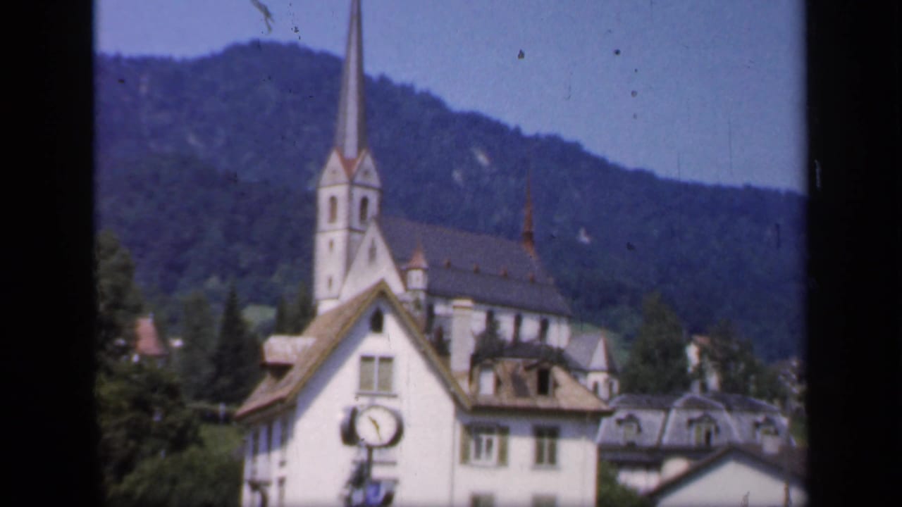 Church spire rising behind a pale building with mountains and trees in the background.
