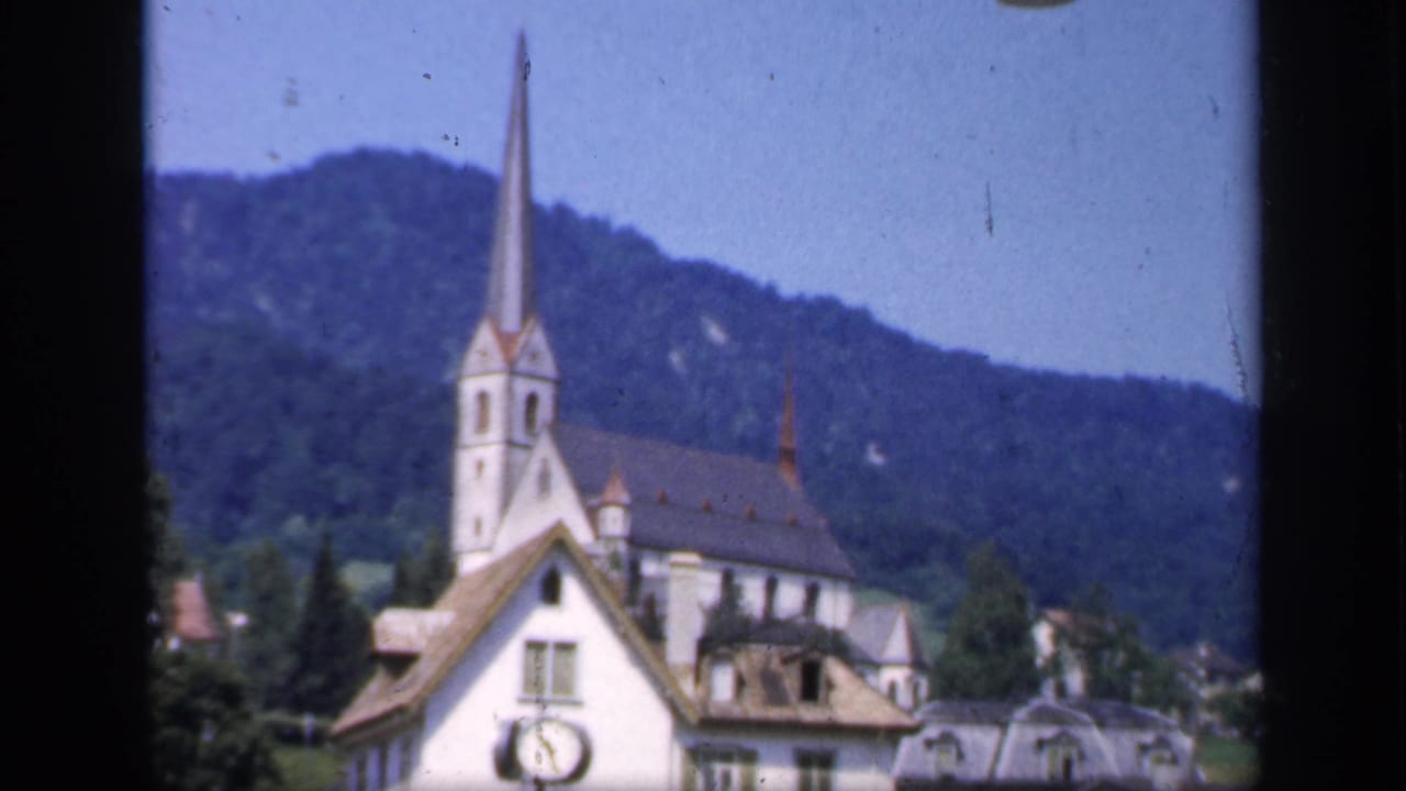 1949 home-movie frame showing the white clock-fronted building, church steeple, nearby roofs, trees, and mountain backdrop.