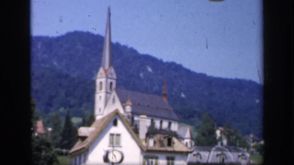 1949 home-movie frame showing the white clock-fronted building, church steeple, nearby roofs, trees, and mountain backdrop.