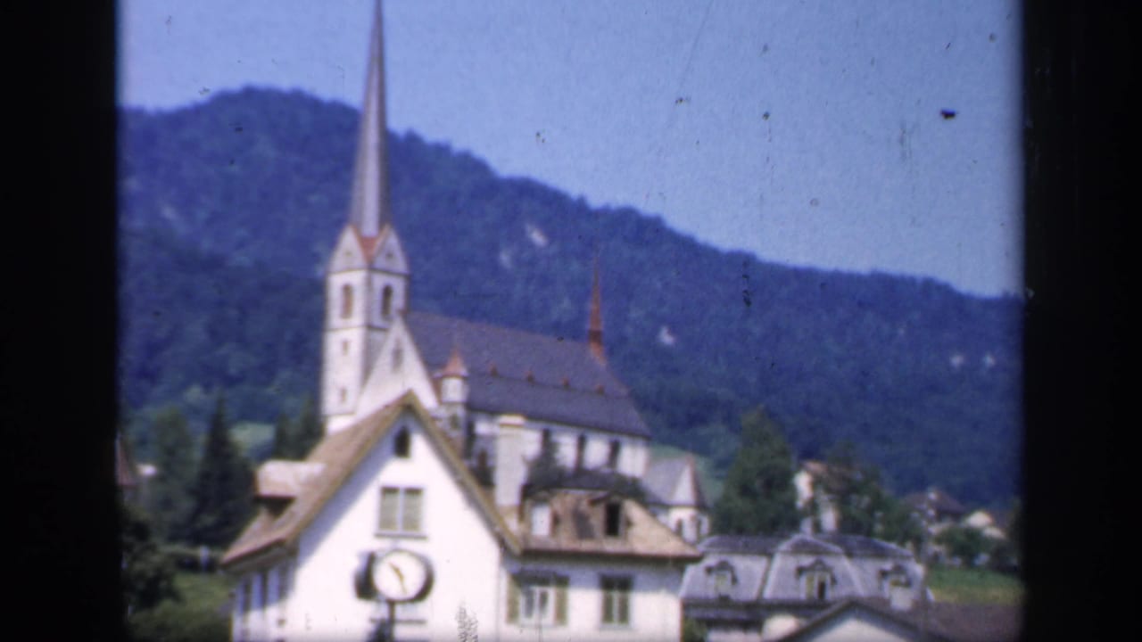 1949 8mm frame of a white church spire rising above village buildings, with wooded hills in the background.