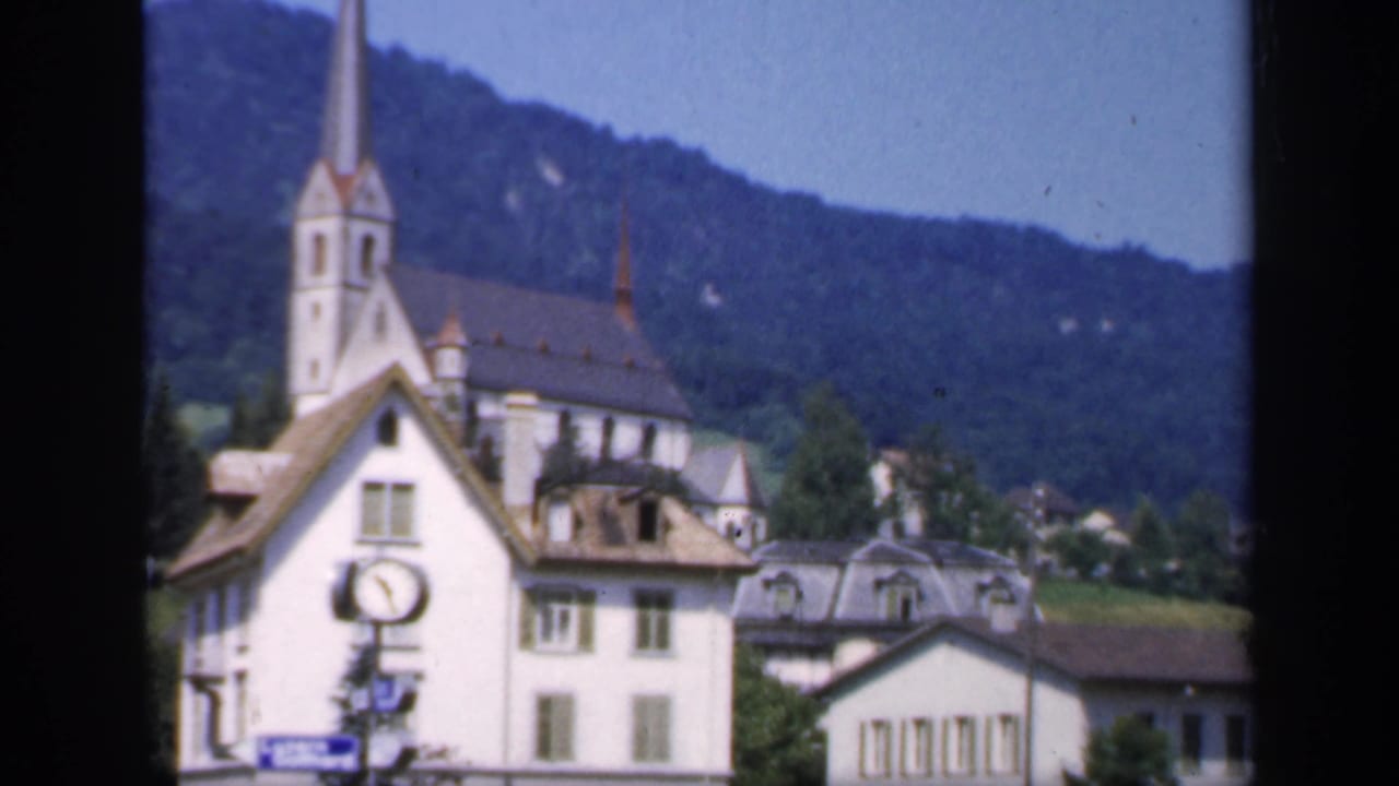 1949 home-movie frame showing a church spire, white buildings, shuttered windows, and a round street clock below wooded hills.