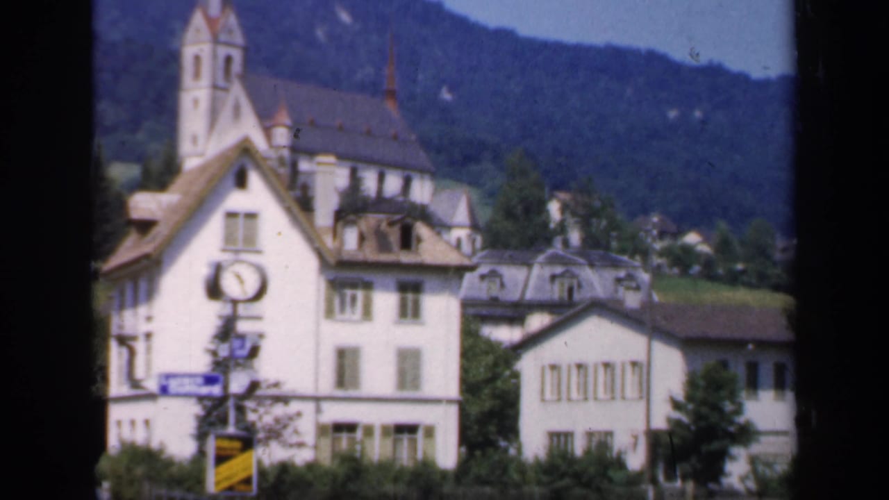 Soft 8mm view of a church spire, white village buildings, a foreground clock, and a green hillside, identified by metadata as England in 1949.