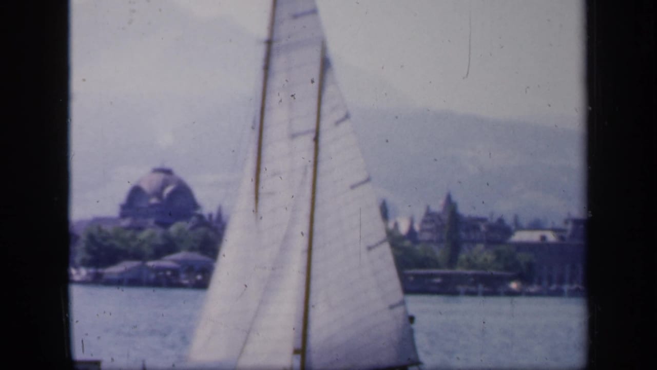 A sailboat passes across the water, with a distant shoreline, trees, and a domed building visible through the soft 8mm frame.