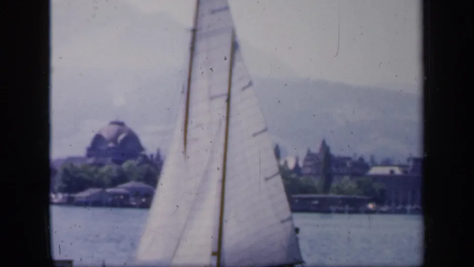 A sailboat passes across the water, with a distant shoreline, trees, and a domed building visible through the soft 8mm frame.