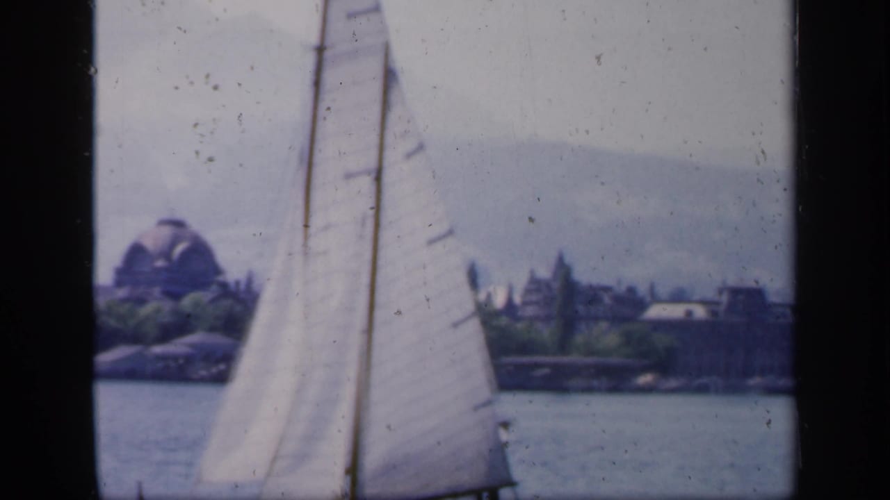 White sailboat passing across calm water with distant shoreline and domed building.