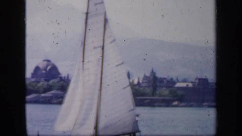 1949 England 8mm frame showing a white sailboat close to camera with a distant lakeside shoreline and domed structure beyond.