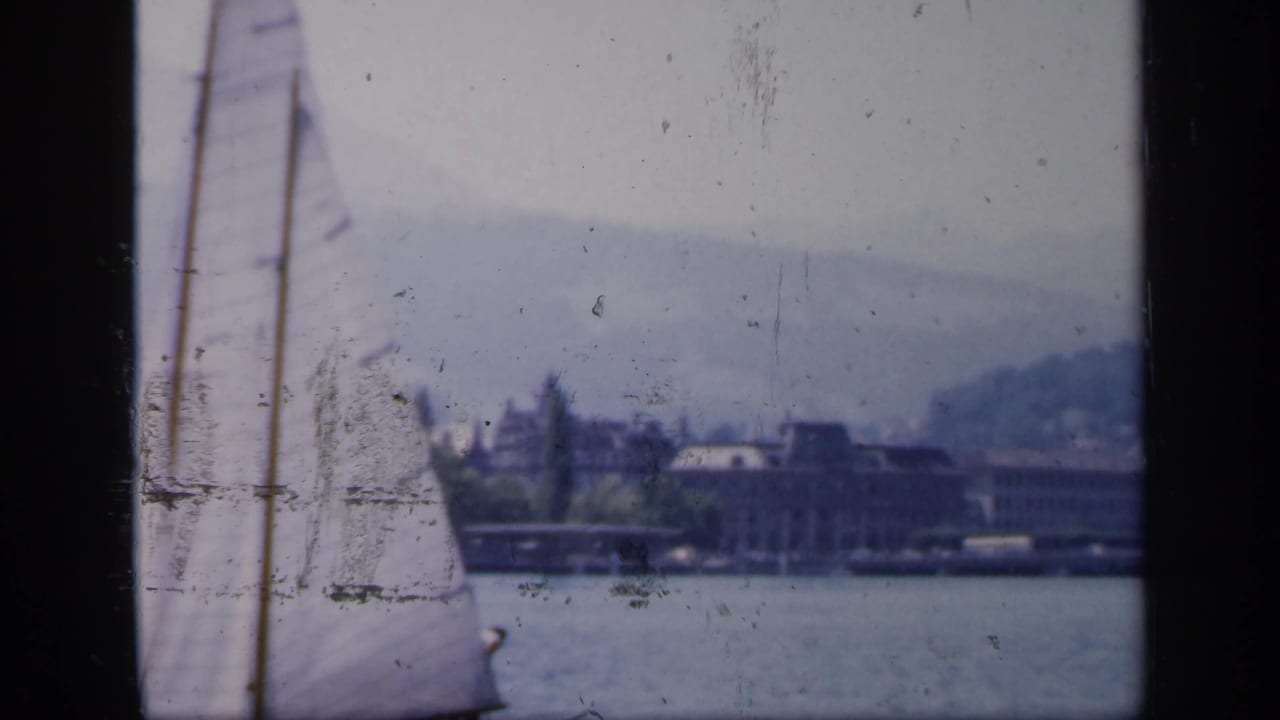 1949 England home-movie frame with a sailboat moving past the lens and waterfront architecture across the lake.