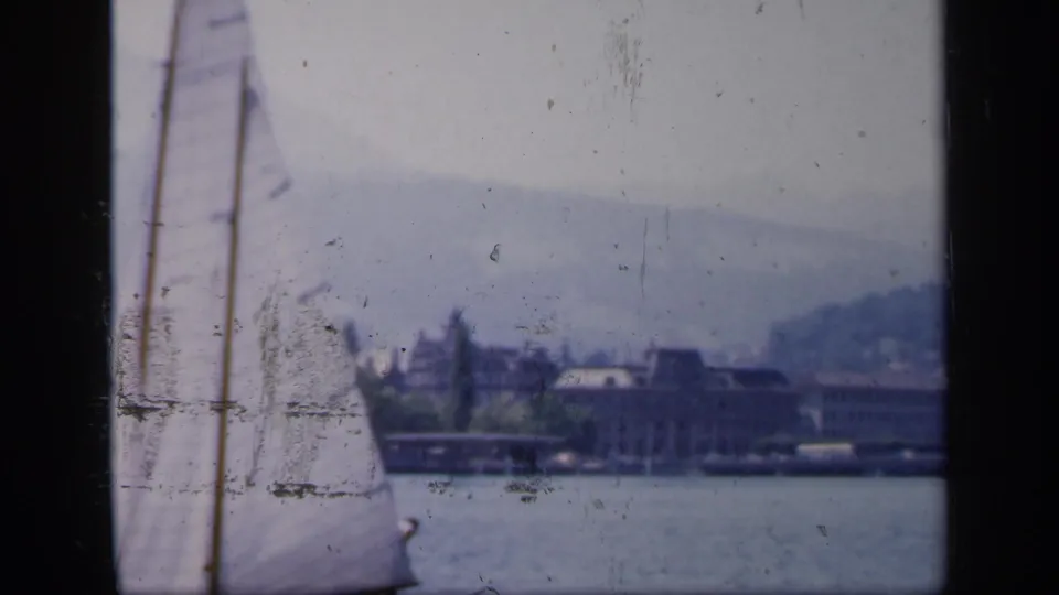 1949 England home-movie frame with a sailboat moving past the lens and waterfront architecture across the lake.