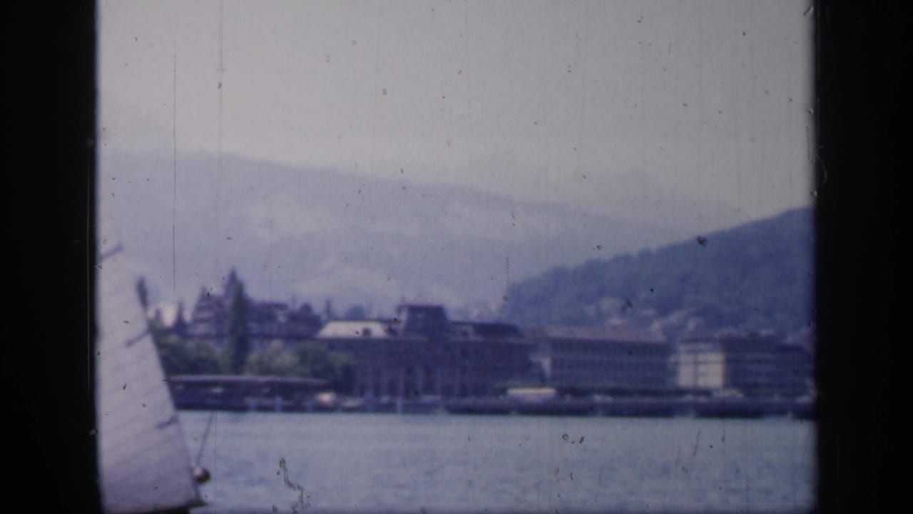 1949 England 8mm view of a partial sail at frame edge, calm water, distant buildings, and a low hillside.