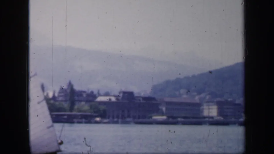 1949 England 8mm view of a partial sail at frame edge, calm water, distant buildings, and a low hillside.