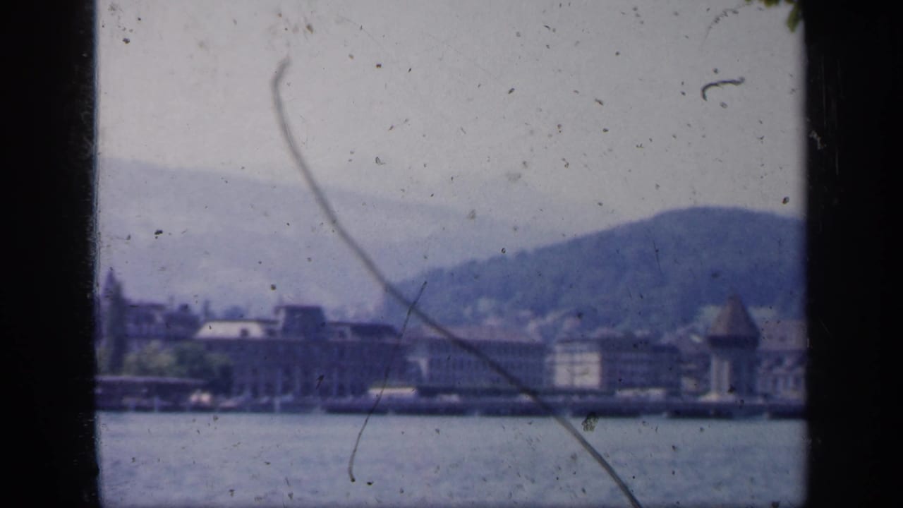 1949 England home-movie frame showing a distant lakeside shoreline and hillside after the sailboat has mostly passed.