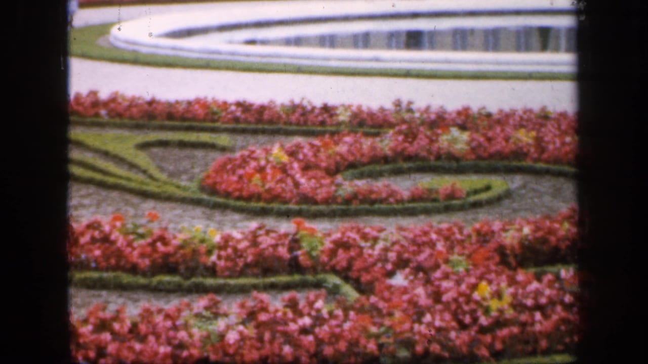 Formal garden beds with pink flowers, clipped hedges, a path, and a fountain in the background.