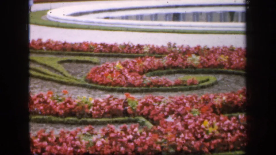 1949 France 8mm frame showing a formal garden with pink flowerbeds, low hedges, pale paths, and part of a circular fountain.