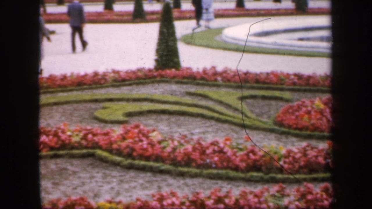 Visitors walk along a pale path between formal flowerbeds and a circular fountain.