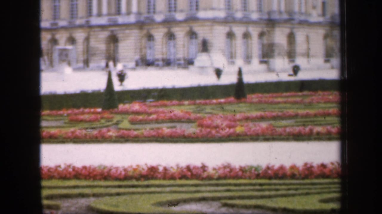 1949 France home-movie frame with a pale palace facade, symmetrical garden beds, red flowers, clipped hedges, and small distant visitors.