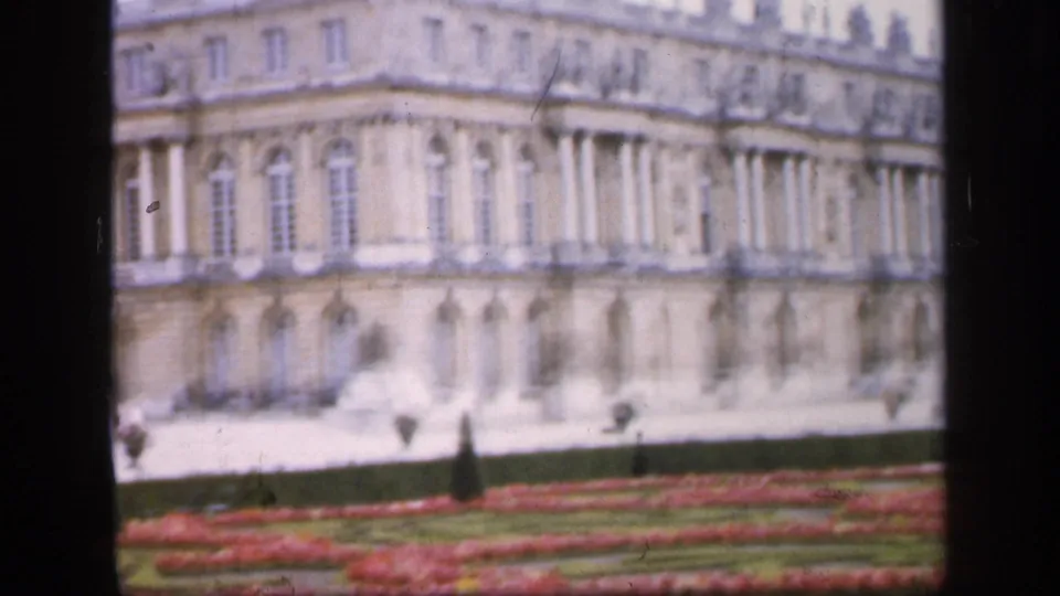 Soft 1949 France frame showing a palace corner with classical columns, arched windows, red flowerbeds, and clipped formal hedges.