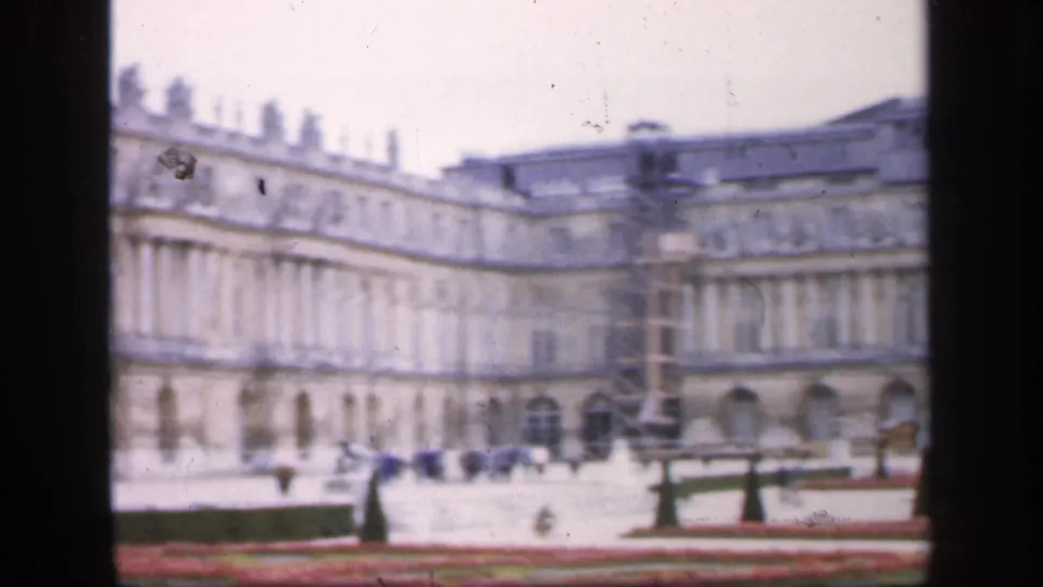 1949 France home-movie frame showing broad palace facades, formal garden beds, paths, and a small distant visitor in the grounds.
