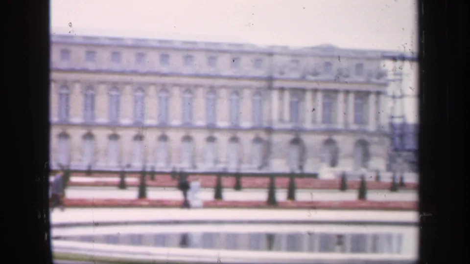 Soft 1949 8mm frame of the Palace of Versailles across a reflecting pool, with garden borders and distant visitors near the facade.
