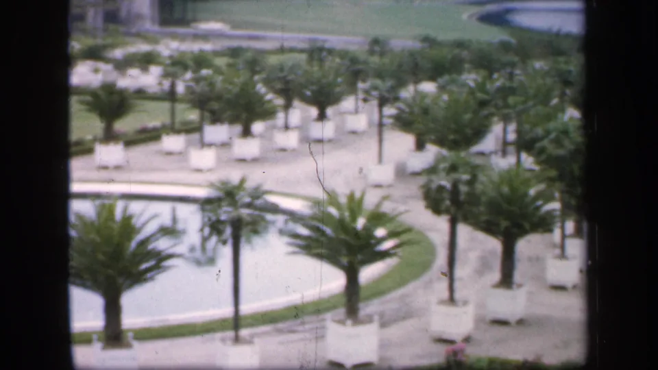 1949 France 8mm frame showing a circular pool in a formal garden, surrounded by palm-like trees, white planters, paths, and trimmed plantings.