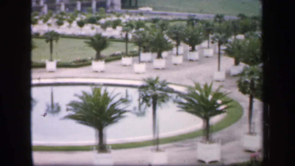 1949 France home-movie frame showing a formal garden through a window, with a circular pool, palms, white planters, paths, and a large building.
