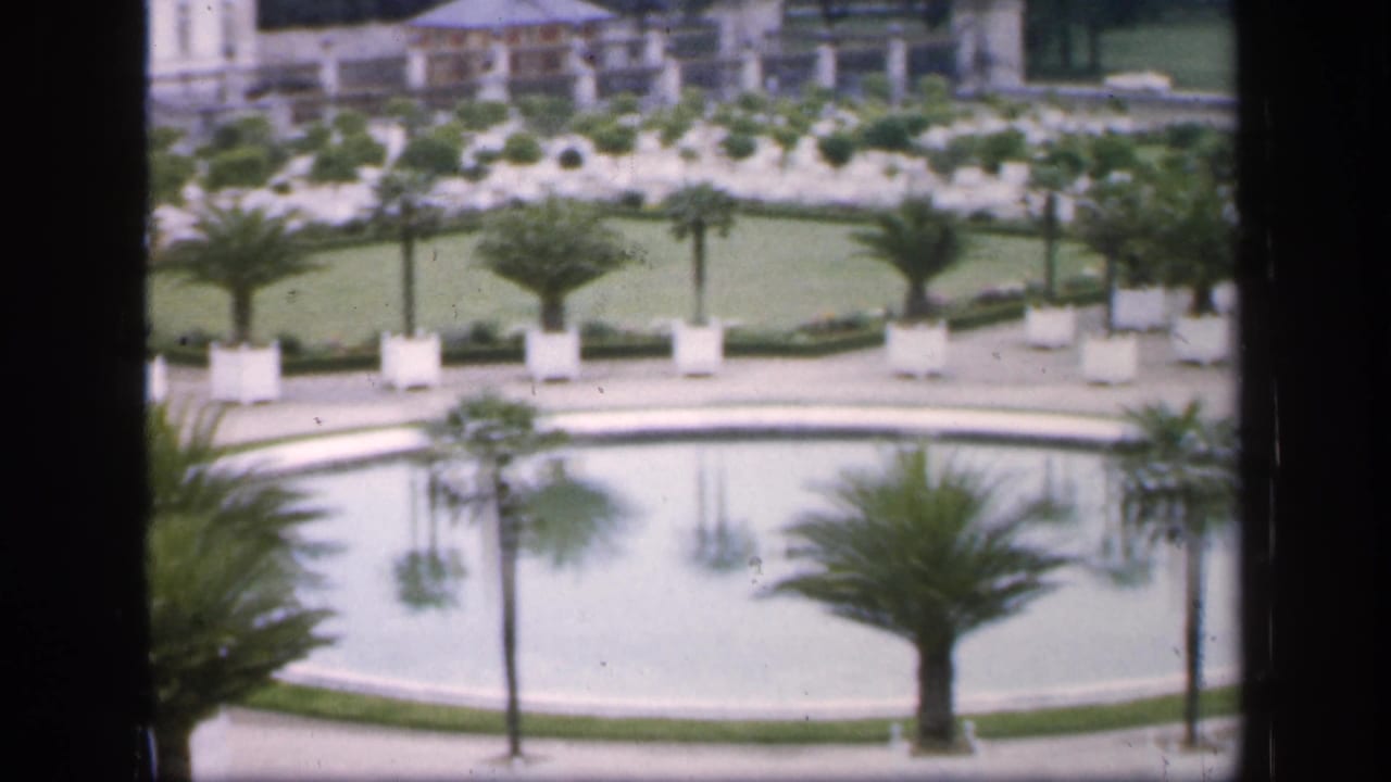 Circular pool and potted palms in a formal garden, partly divided by a dark vertical window frame.